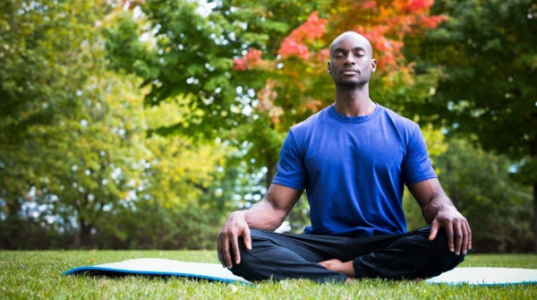 Man in a blue t-shirt sitting cross-legged on a yoga mat at a park, eyes closed