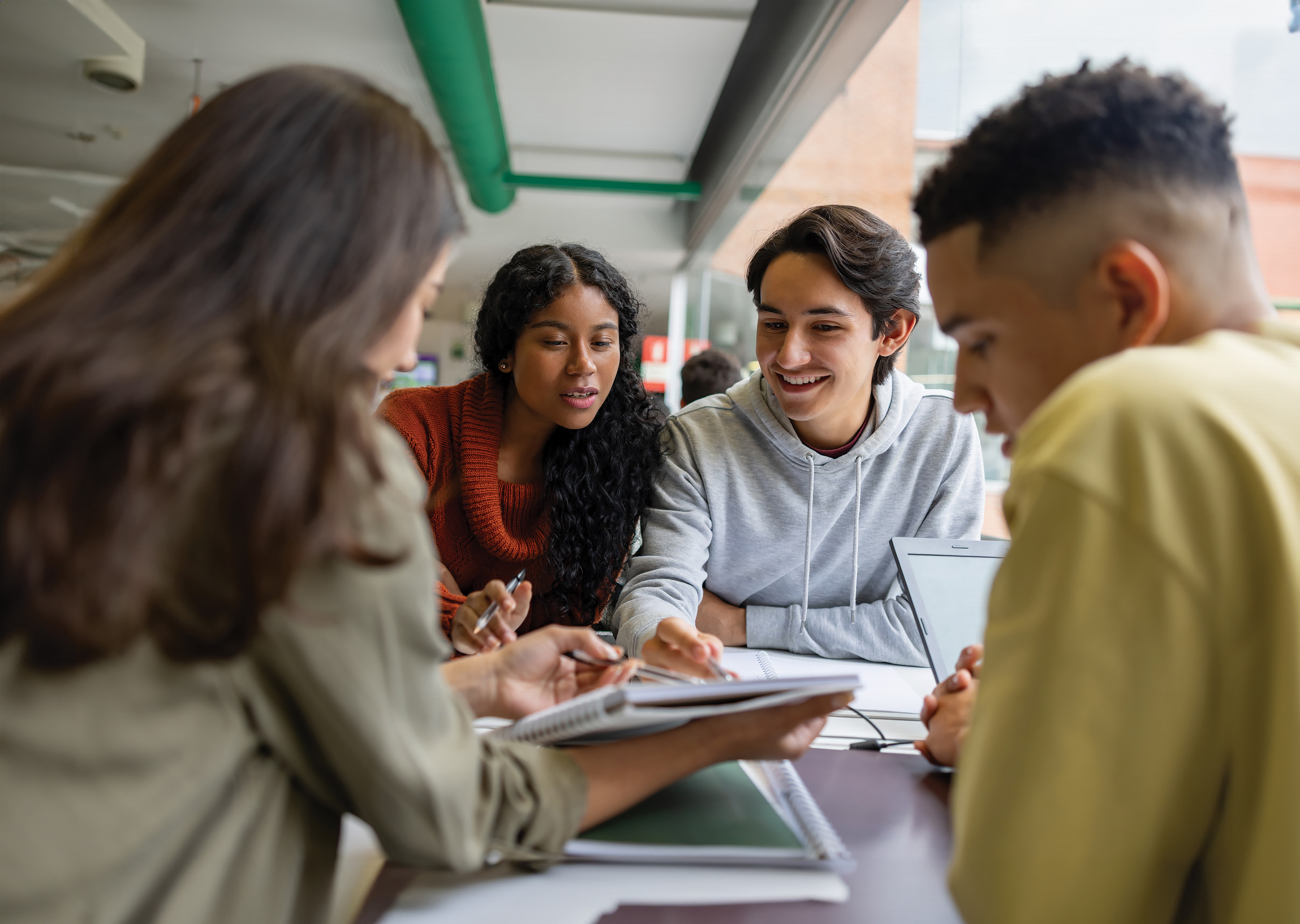 Four diverse college students collaborate around a table with notebooks, smiling and discussing schoolwork together.