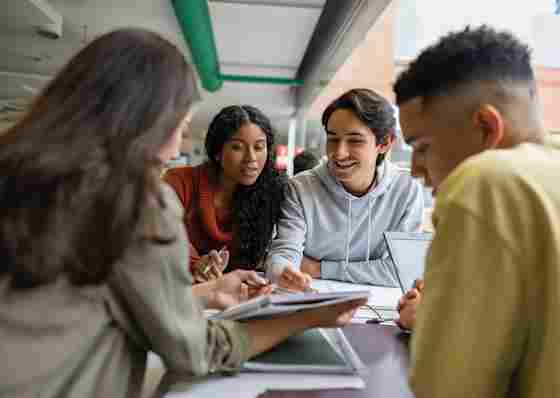 Four diverse college students collaborate around a table with notebooks, smiling and discussing schoolwork together.