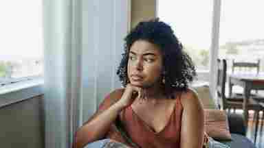 A young woman looks pensively out a window in a quiet, sunlit room.