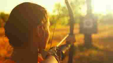 A woman standing in a field aims an arrow at her target at sunset.