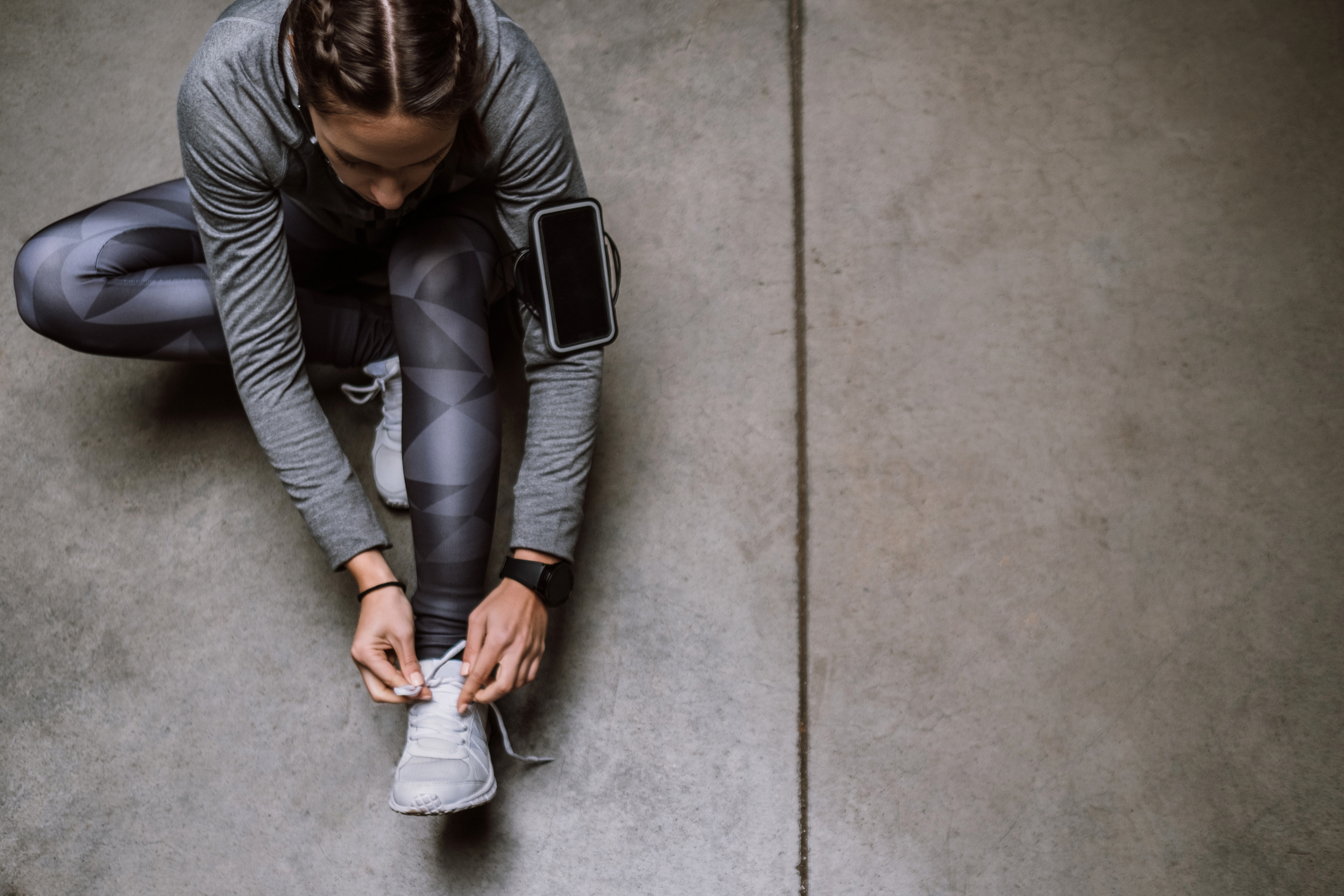 A woman in gray athletic wear ties her white sneakers on a concrete floor, with a phone armband and fitness tracker visible.