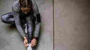 A woman in gray athletic wear ties her white sneakers on a concrete floor, with a phone armband and fitness tracker visible.