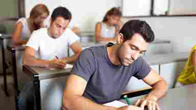 A focused male adult student is writing in a classroom setting with other students in the background. College. Young adult. Man.