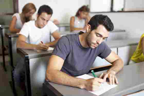 A focused male adult student is writing in a classroom setting with other students in the background. College. Young adult. Man. 