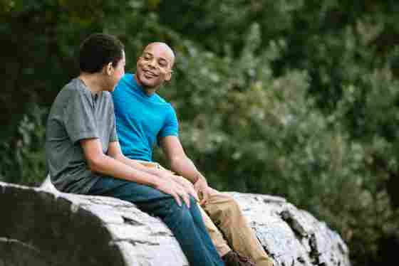 A man talks to his son while sitting on a fallen tree, enjoying the outdoors.