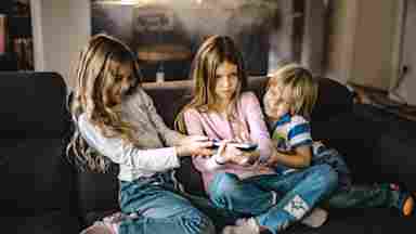 Three young children on a living room sofa squabbling over a remote control while watching TV.