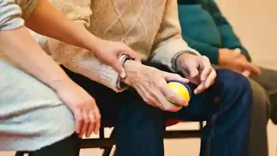 A caregiver gently holds an elderly person's wrist as they grip a small yellow and blue ball during a therapy session.