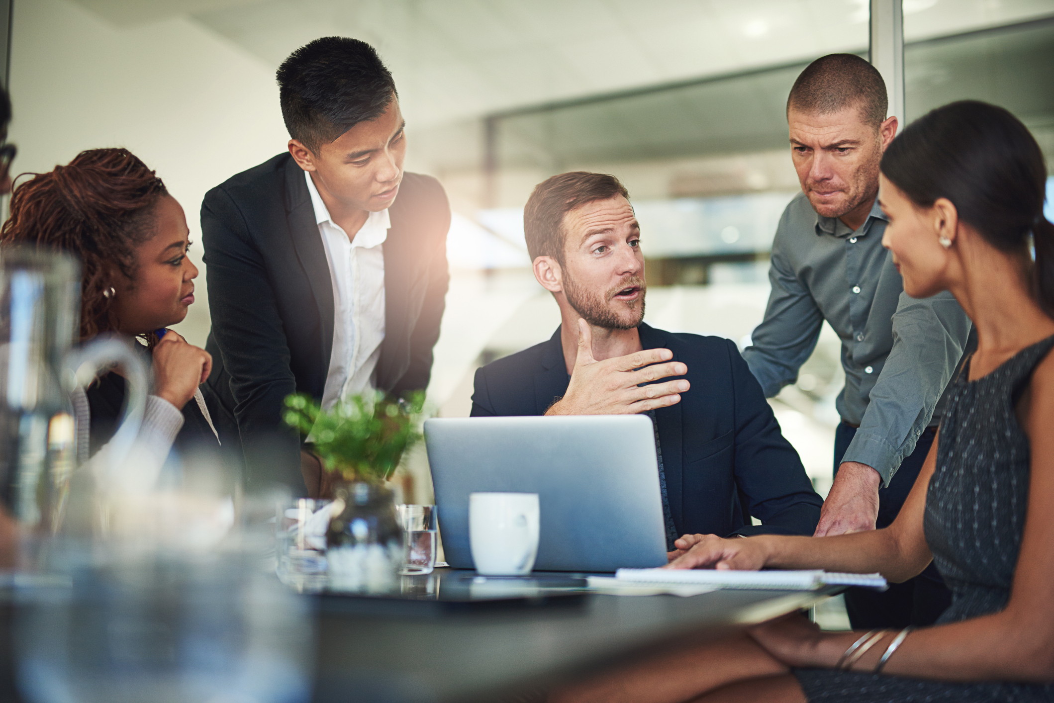 A group of colleagues have a dynamic exchange in a sunlit board room.