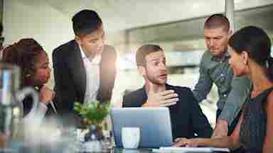 A group of colleagues have a dynamic exchange in a sunlit board room.