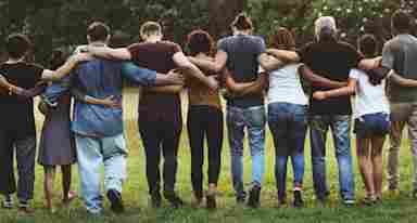 A diverse group of adults standing outdoors with their backs to the viewer and their arms around each other's shoulders.