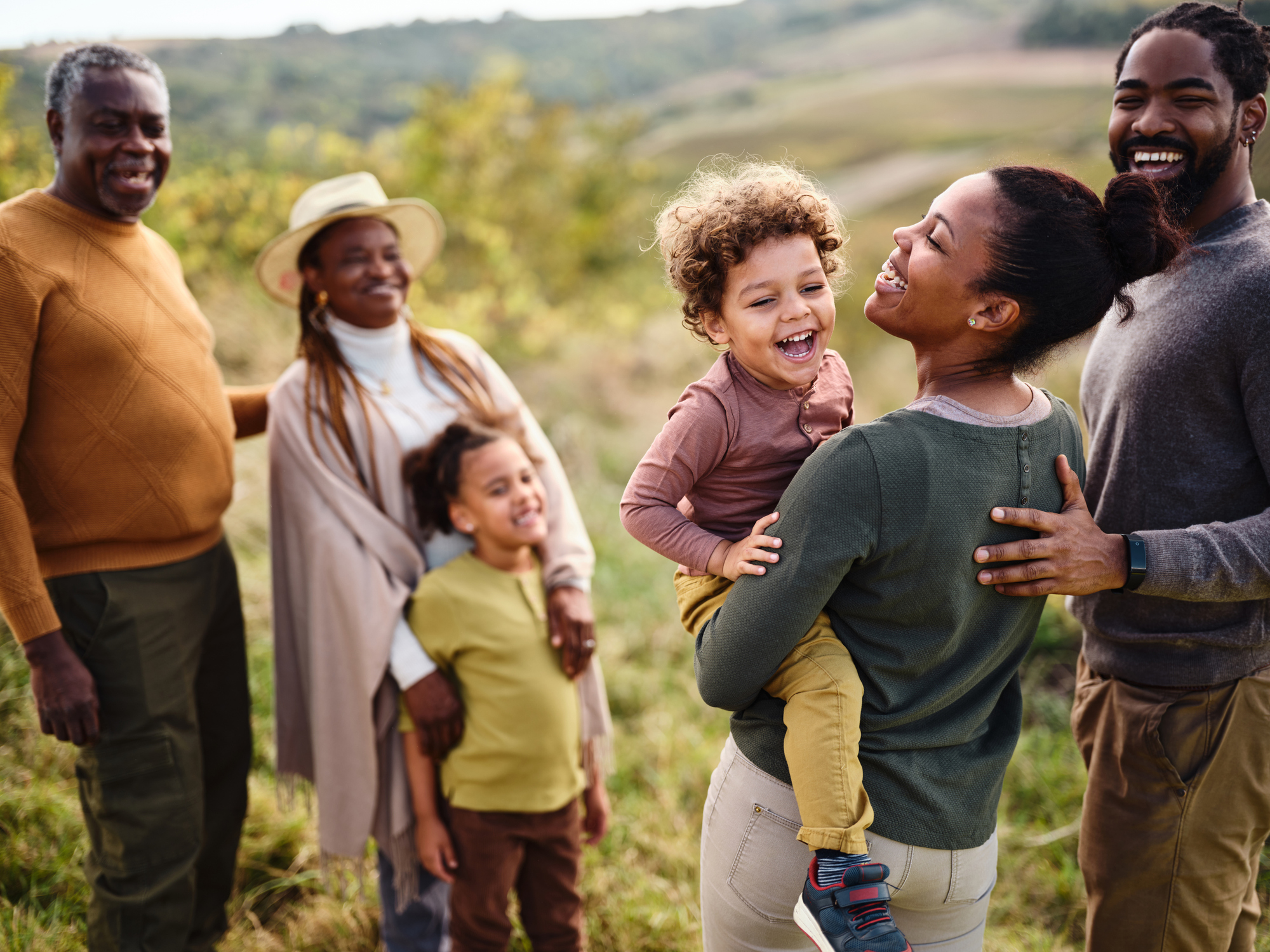 A joyful multigenerational family laughing outdoors in a green field, with a mother lifting a young child while grandparents smile nearby.