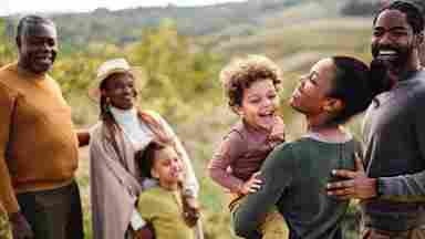 A joyful multigenerational family laughing outdoors in a green field, with a mother lifting a young child while grandparents smile nearby.