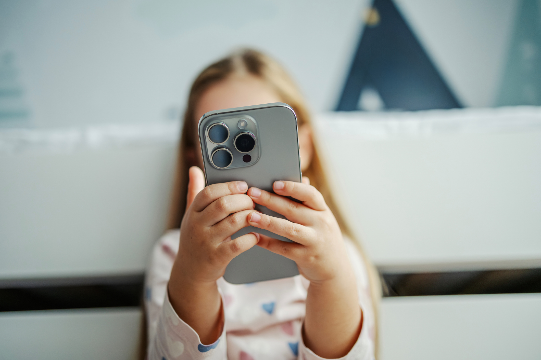 A young blonde girl holds a gray smartphone up to her face, obscuring it, while sitting on a white couch.