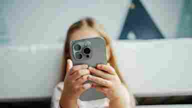 A young blonde girl holds a gray smartphone up to her face, obscuring it, while sitting on a white couch.