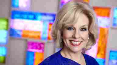 A headshot of Dr. Sandra Bond Chapman standing in front of a multicolored background. She is wearing a dark blue blouse and smiling at the viewer.