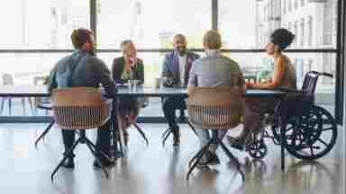 A woman in a wheelchair meets with her team in a sunlit room to discuss strategy.