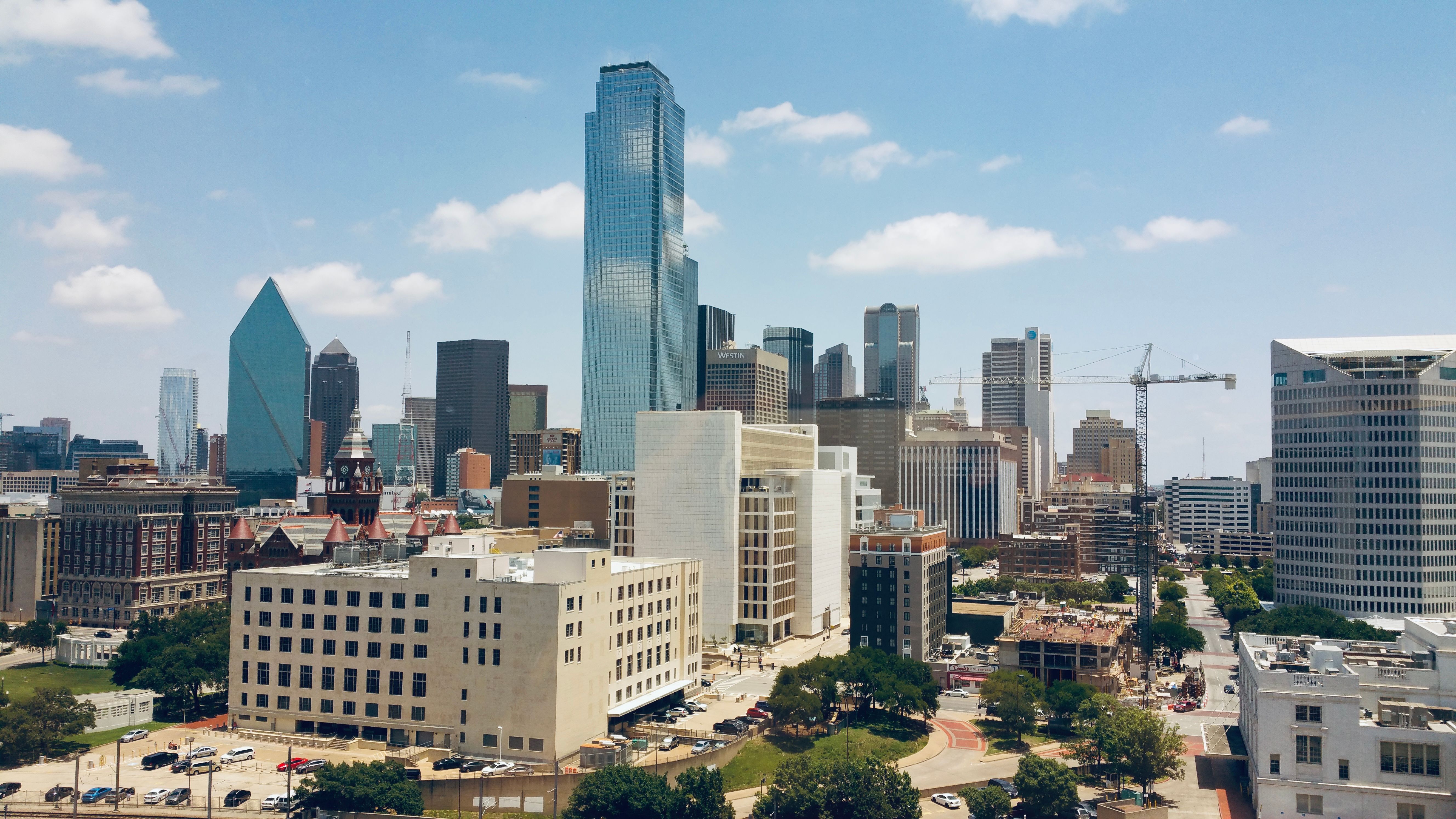Aerial view of Dallas, Texas skyline featuring glass skyscrapers, historic buildings, construction cranes, and a partly cloudy blue sky. Photographer: Anat Morad.