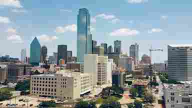 Aerial view of Dallas, Texas skyline featuring glass skyscrapers, historic buildings, construction cranes, and a partly cloudy blue sky. Photographer: Anat Morad.