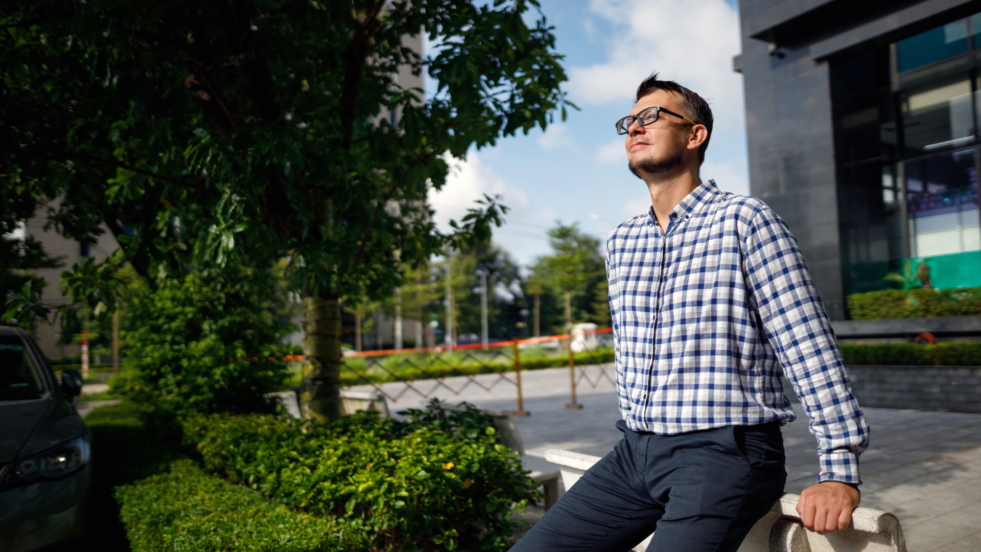 A young professional in plaid shirt gazes across the lawn outside an off building, taking a break in some brain-healthy sunshine. 