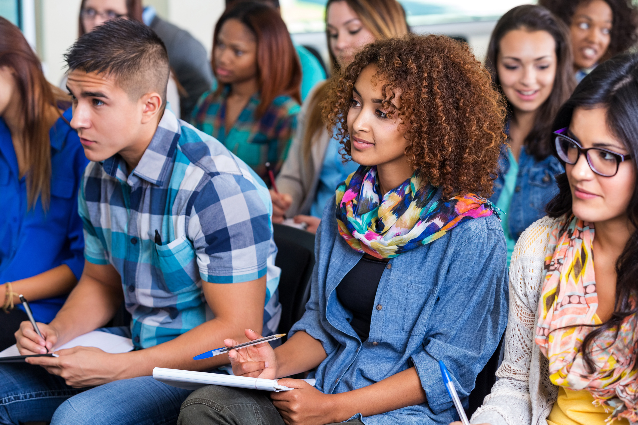 A large, diverse group of teens takes notes during a lecture.