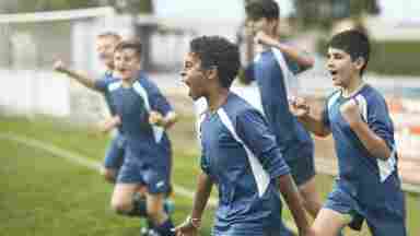 Energetic preteen/teenage male footballers cheering and punching the air as they run onto the field for a practice session.