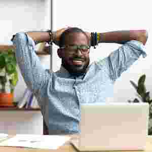 A happy man is on his computer with his hands behind his head.