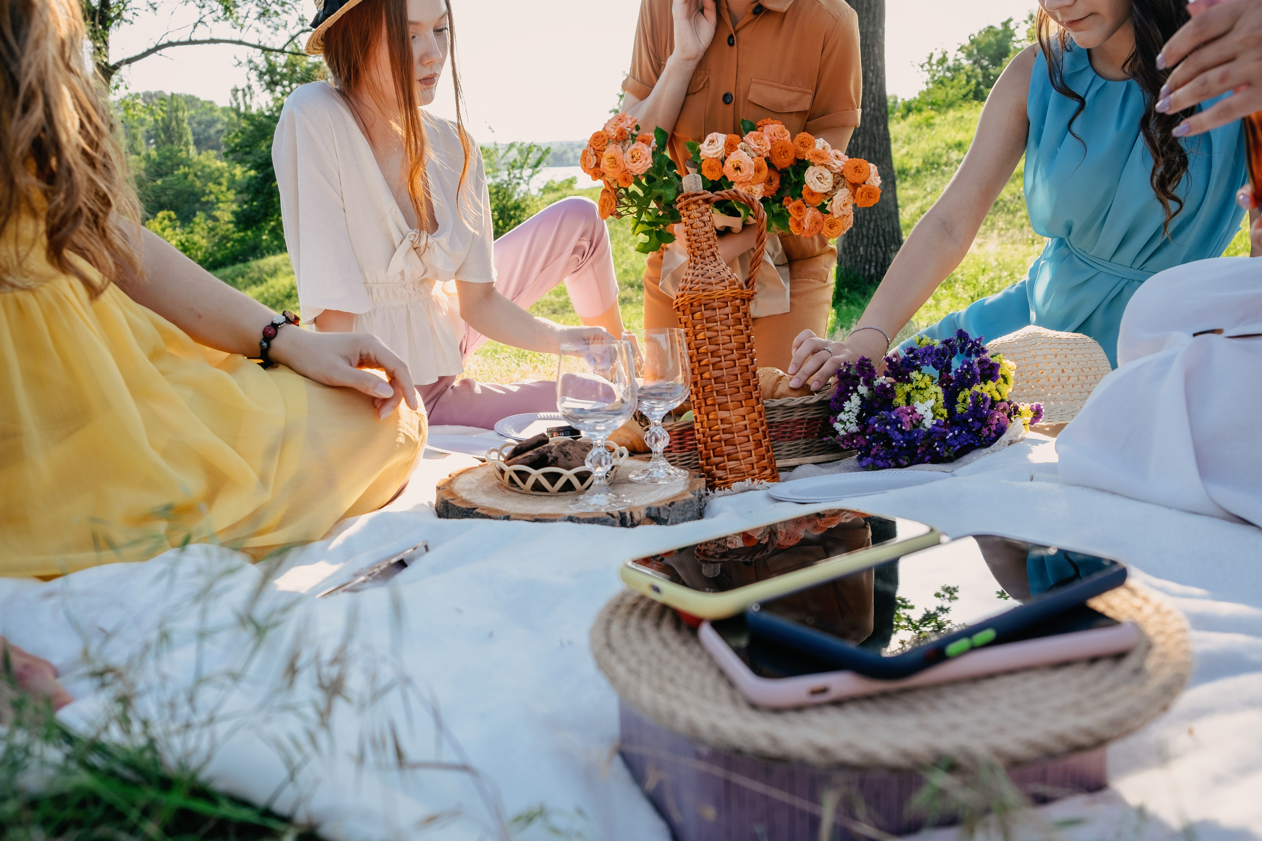 Women gathered around a picnic blanket outdoors with flowers, a wicker bottle, wine glasses, food, and phones on a sunny day.