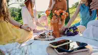 Women gathered around a picnic blanket outdoors with flowers, a wicker bottle, wine glasses, food, and phones on a sunny day.