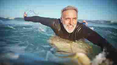 A smiling middle-aged man with gray hair and beard in a black wetsuit paddling on a surfboard in ocean waves.