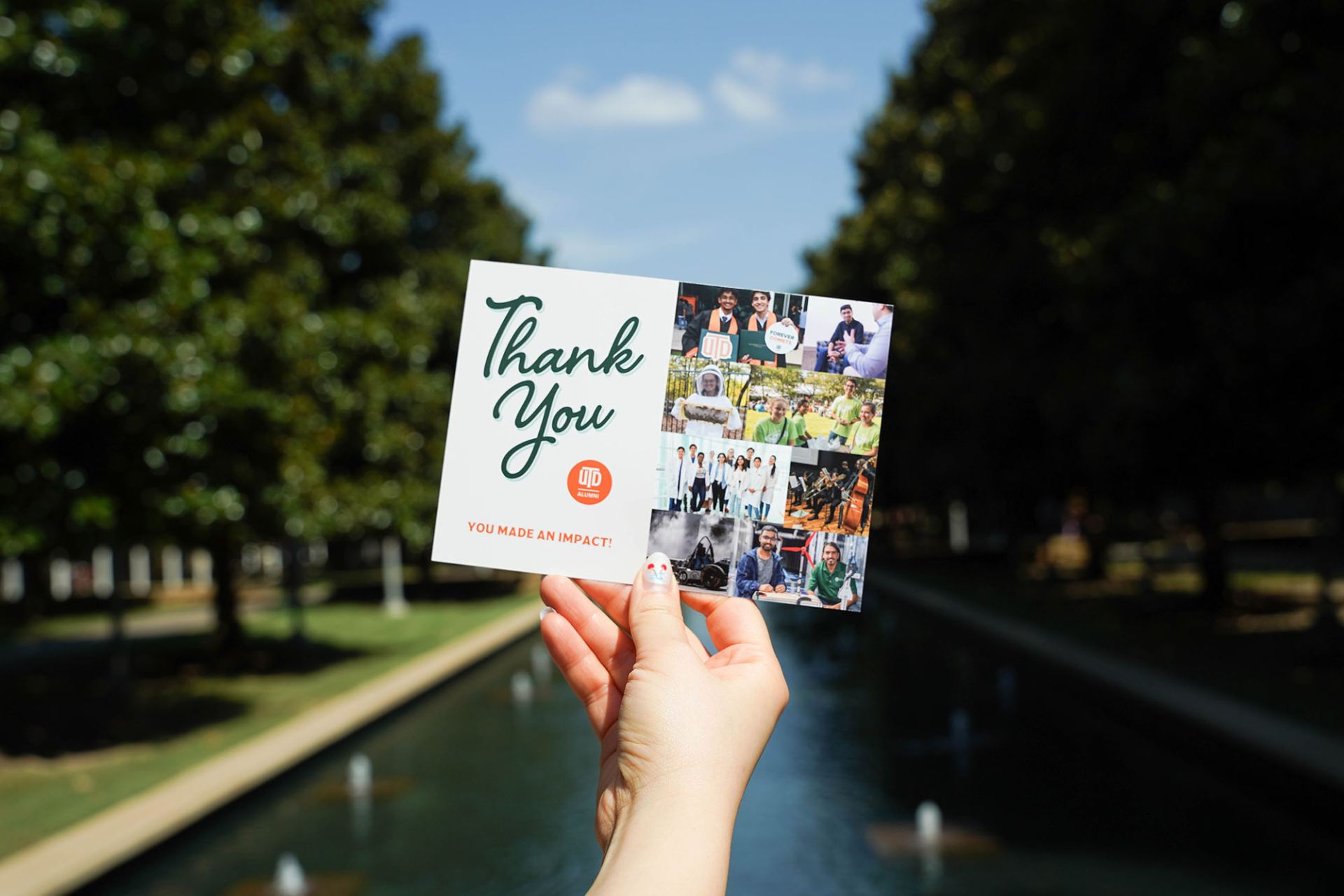 A hand holds a UTD Alumni "Thank You, You Made an Impact!" card with a photo collage outdoors near a fountain.