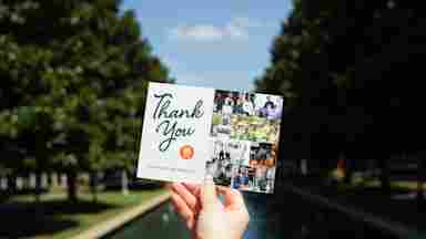 A hand holds a UTD Alumni "Thank You, You Made an Impact!" card with a photo collage outdoors near a fountain.