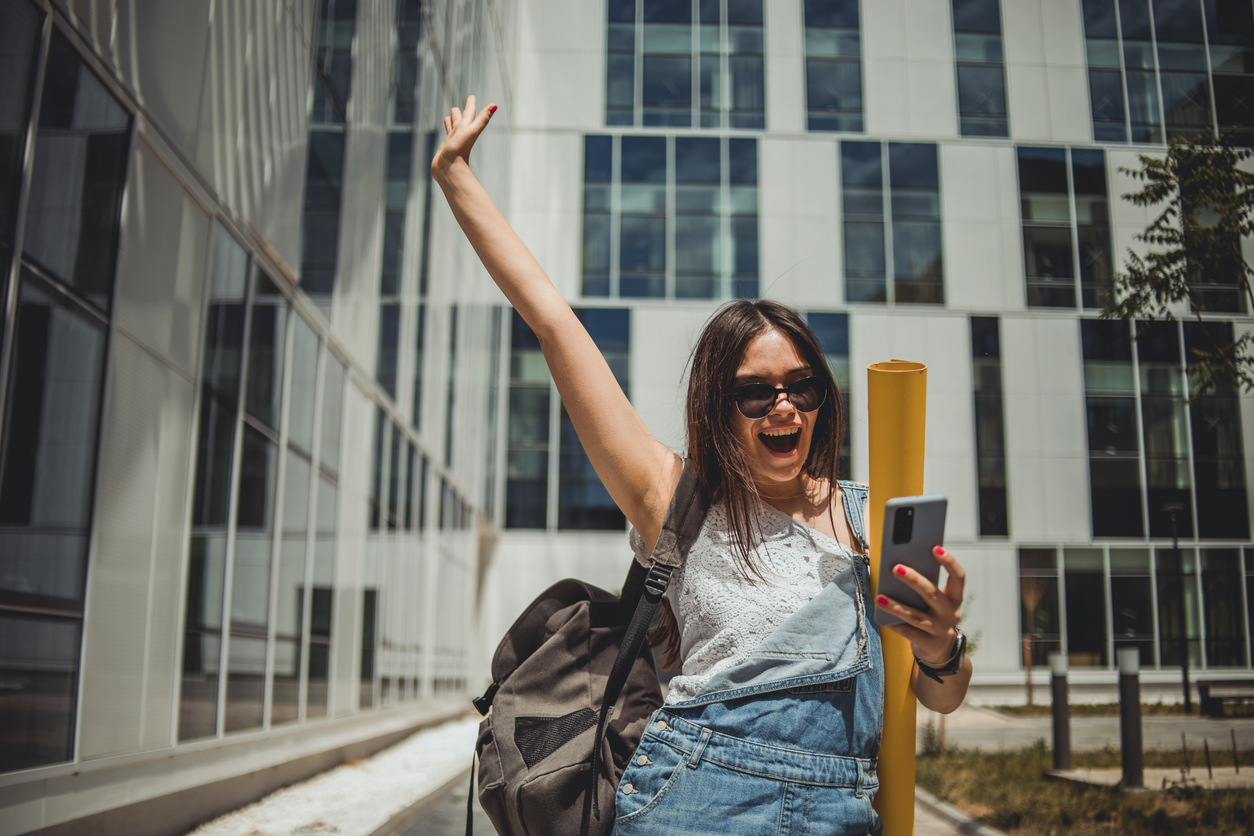 Joyous college student standing outside campus buildings as she looks at her grades.