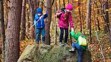 Children with backpacks hiking in autumn forest and helping one another climb a large rock.