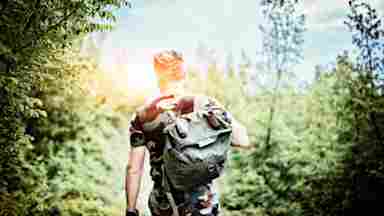 A soldier walks alone in a mountain forest, carrying a backpack.