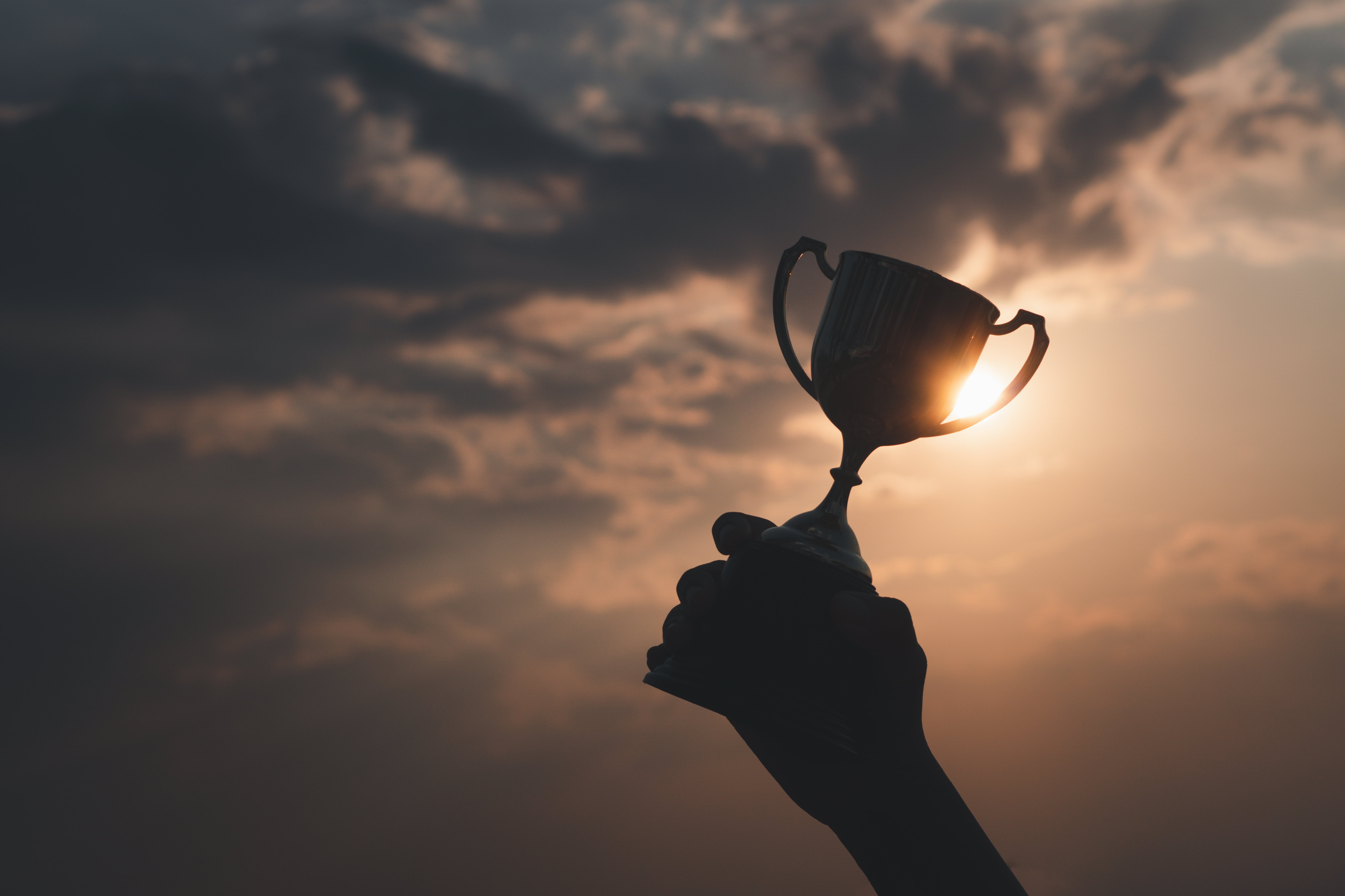 Silhouette of a hand raising a trophy cup against a dramatic sunset sky with glowing clouds.