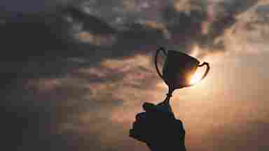 Silhouette of a hand raising a trophy cup against a dramatic sunset sky with glowing clouds.