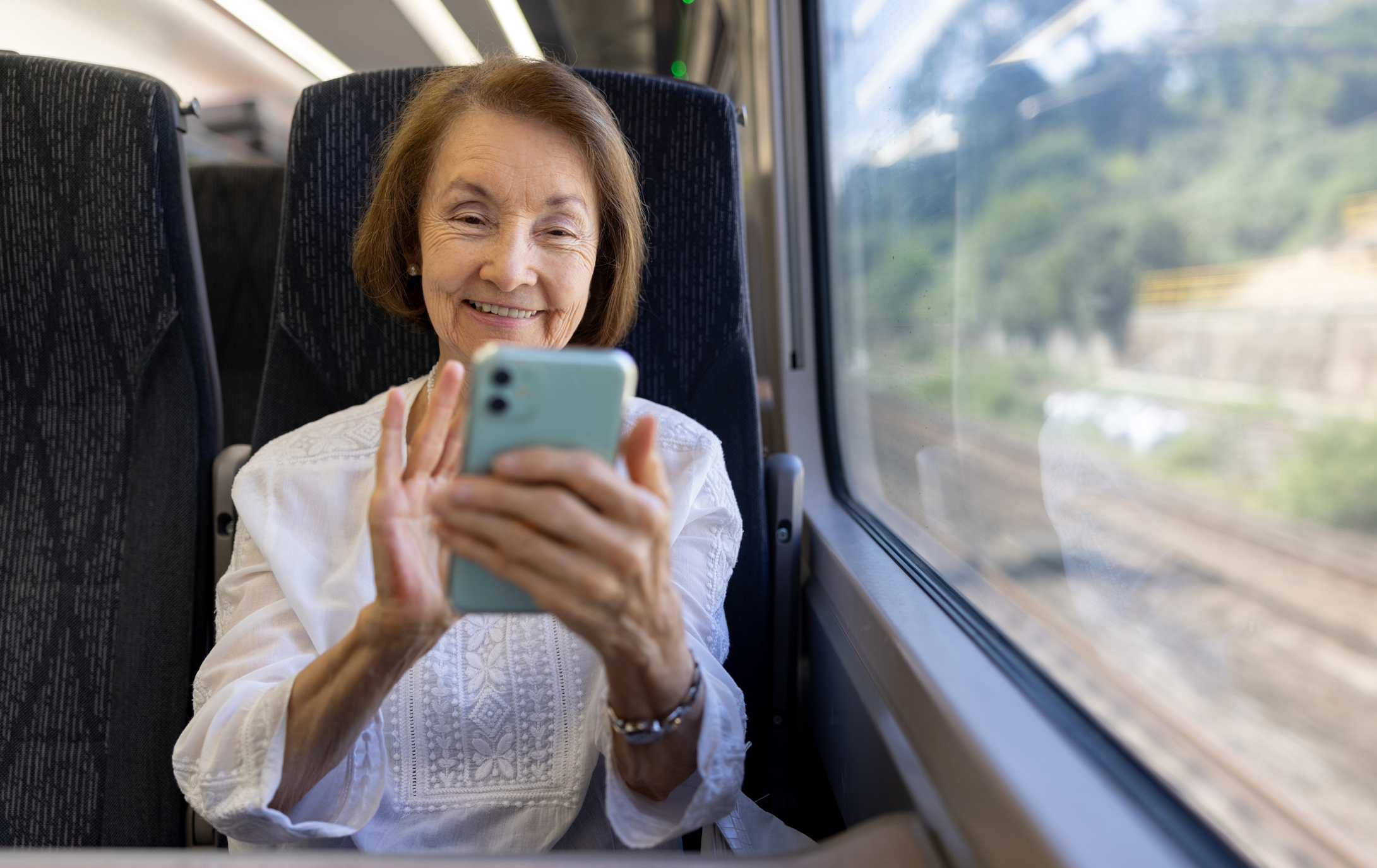An older woman smiles while using a smartphone on a train, wearing a white blouse, with greenery visible through the window.