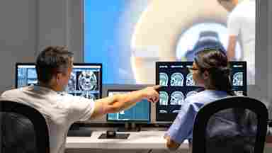 Researchers analyze MRI scan results, while a study participant receives a scan in the background.
