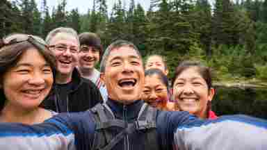 A group of related backpackers smile look at a mobile phone while taking a selfie near a lake in a wilderness park. The group is a real multi-ethnic and multi-generation extended family of mature adults and teenage children. Wide angle view of the forest and pond in the background.