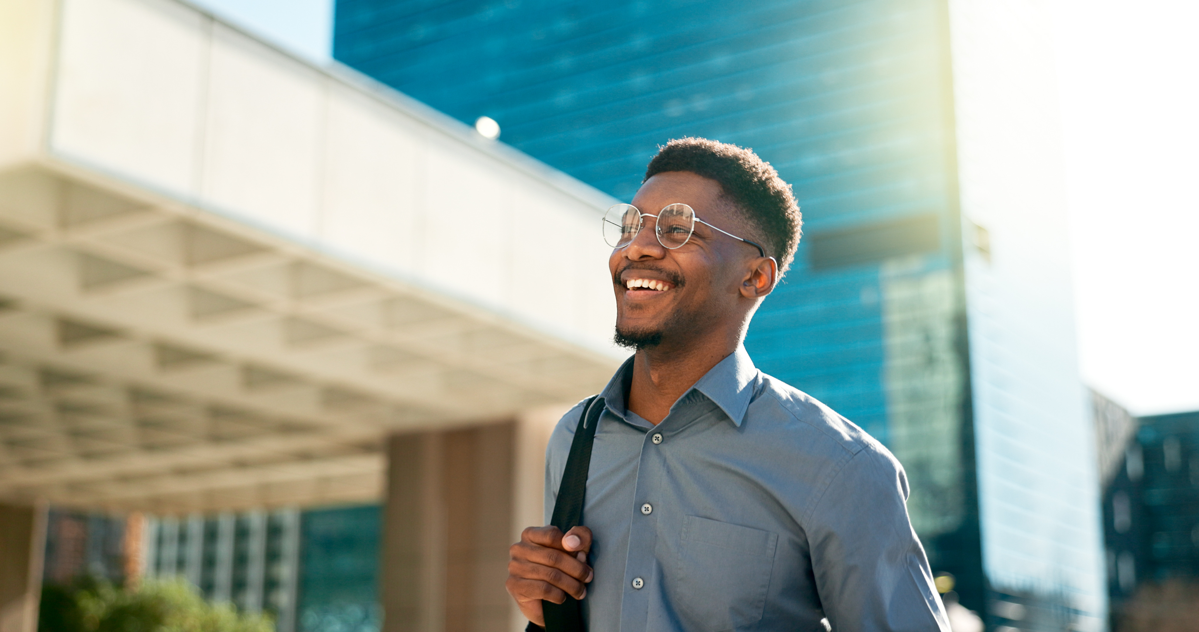 A man walking outside and smiling in the sun. He is dressed in office attire.