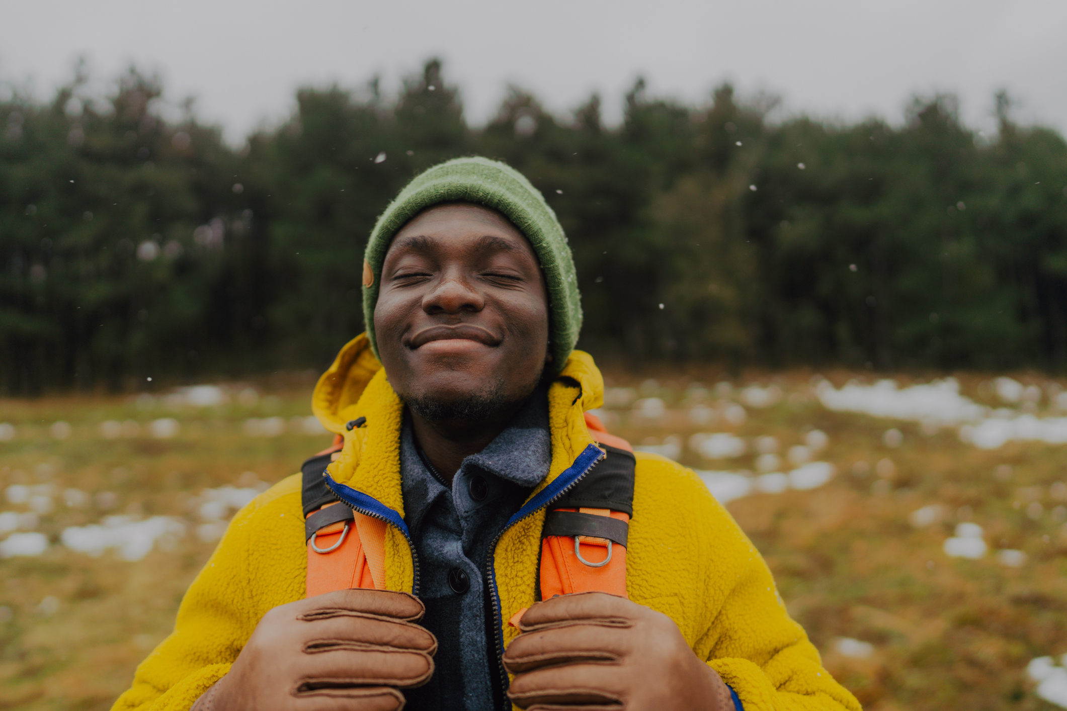A Black man smiles in a yellow fleece jacket, green beanie, and brown gloves, standing outdoors in a snowy field with trees behind him.