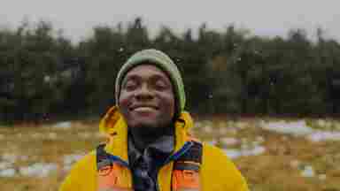 A Black man smiles in a yellow fleece jacket, green beanie, and brown gloves, standing outdoors in a snowy field with trees behind him.