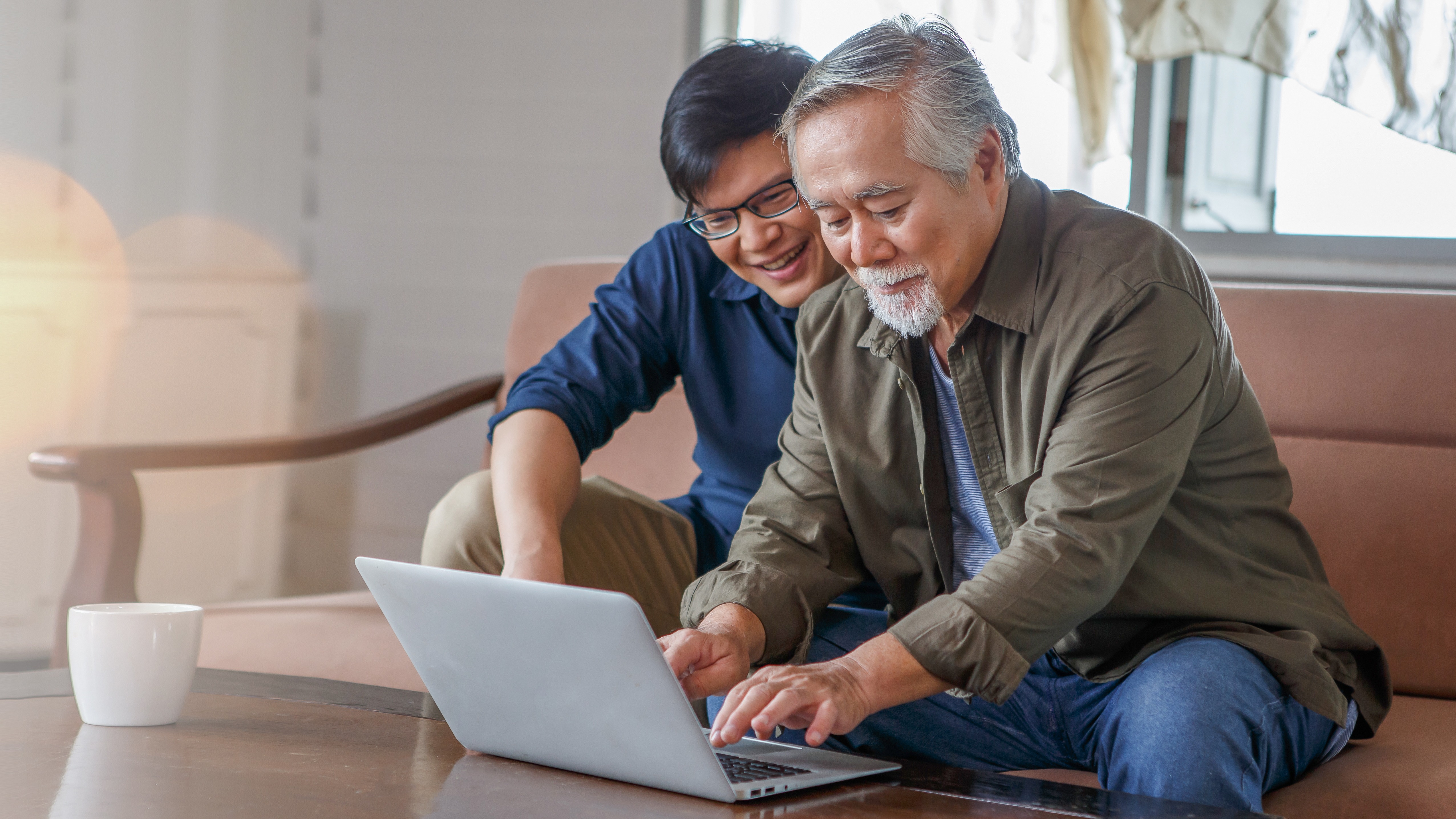 A young man smiling and pointing at a laptop screen while an older man types, both seated together in a cozy living room.