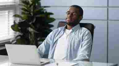 A relaxed man sits in front of computer with eyes closed. This setting is in an office. 