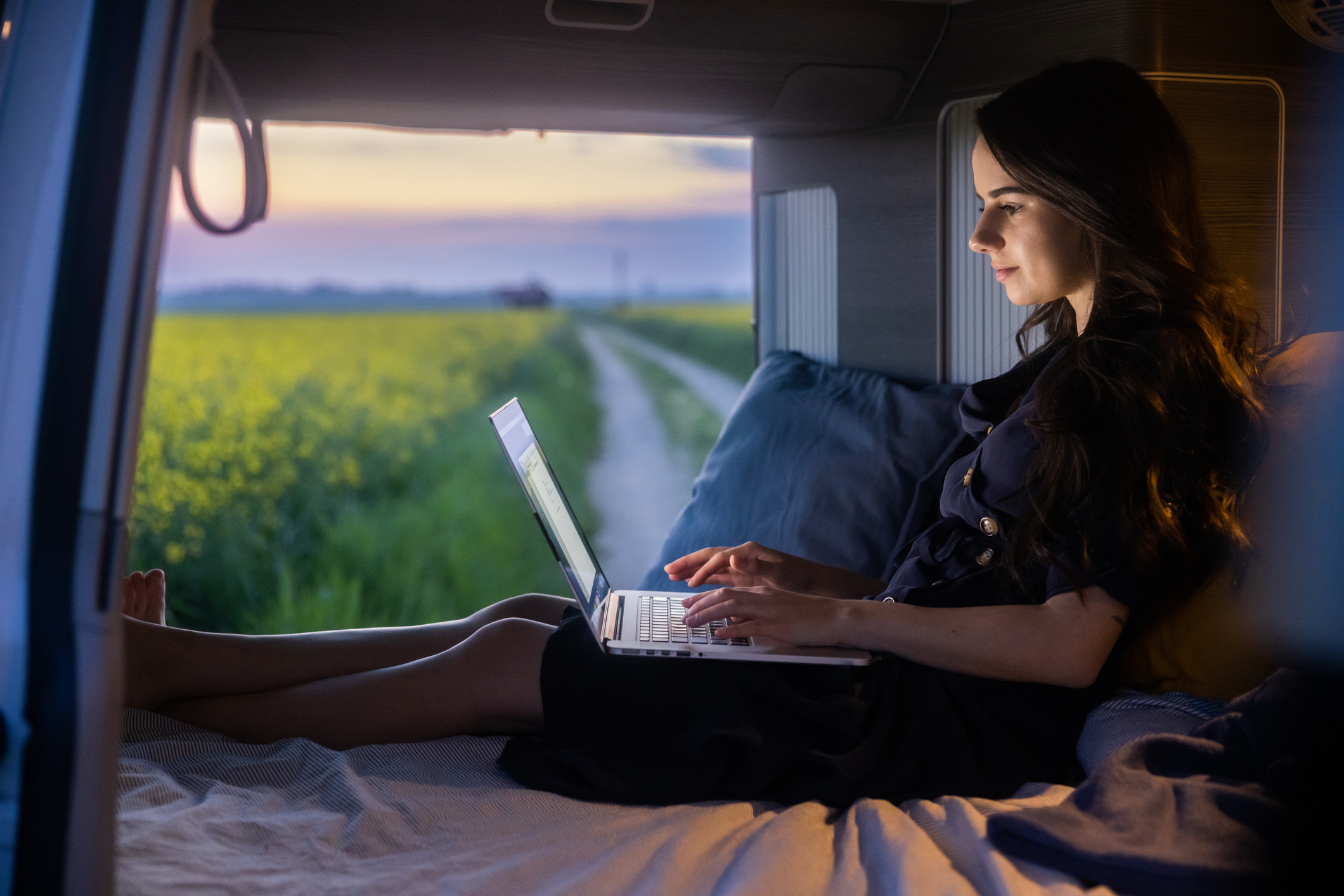 Young person sits on a mattress inside a travel van, working on a laptop beside a beautiful field of flowers.