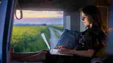 Young person sits on a mattress inside a travel van, working on a laptop beside a beautiful field of flowers.