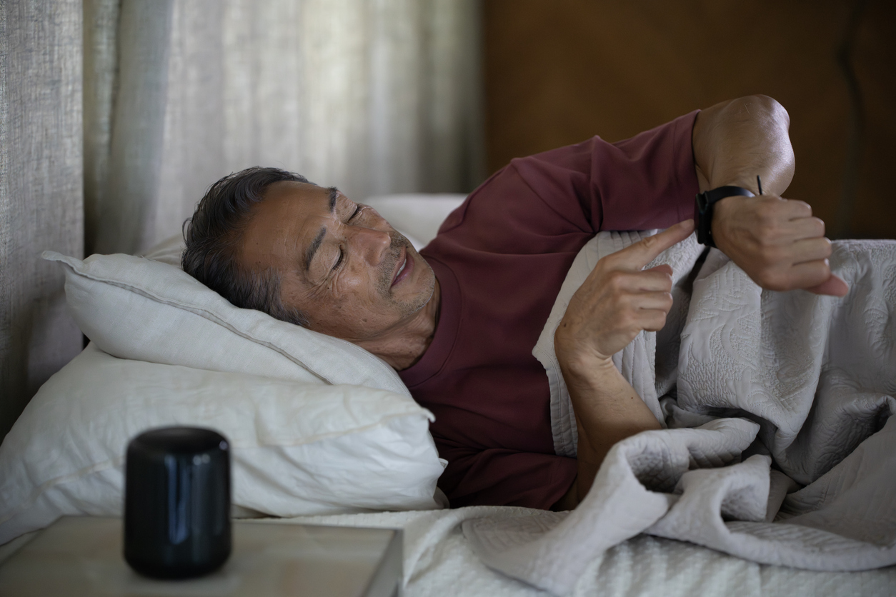 Person in burgundy shirt lying in bed checking smartwatch, with a black speaker on bedside table.