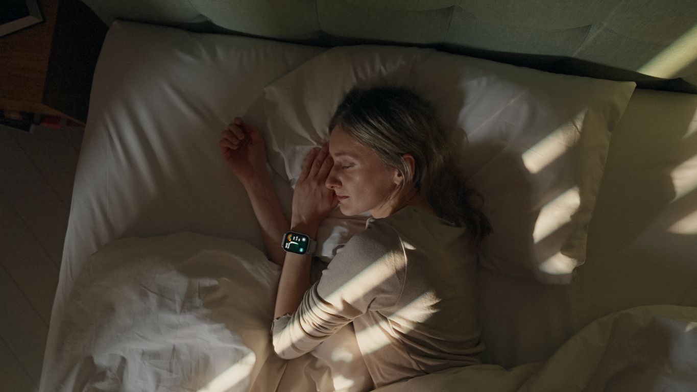 Person sleeping peacefully in bed with white sheets, wearing a smartwatch, as sunlight streams through window blinds.