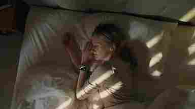 Person sleeping peacefully in bed with white sheets, wearing a smartwatch, as sunlight streams through window blinds.